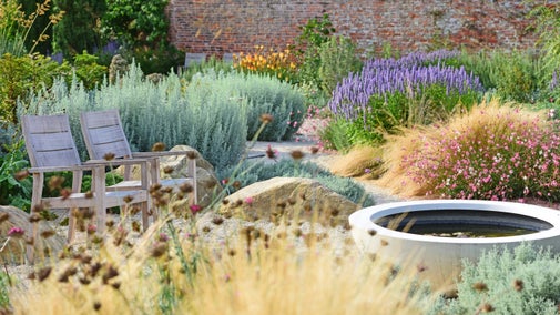 Two wooden chairs in a gravel garden surrounded by plants and grasses with a large round water bowl